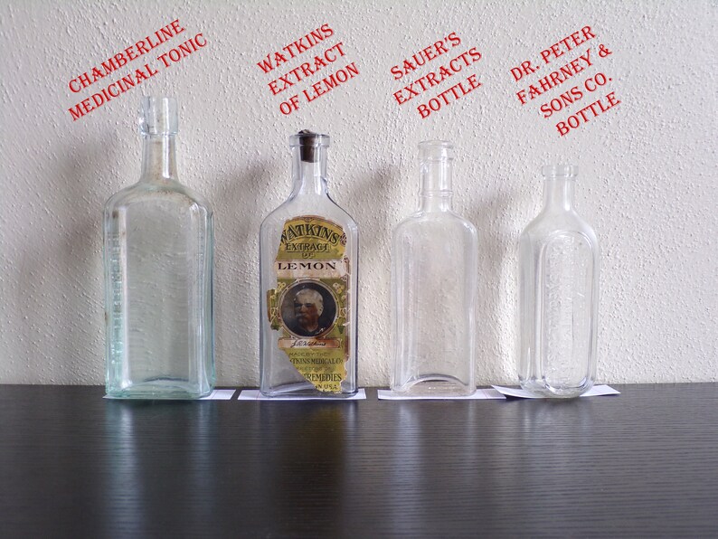 May include: Four clear glass bottles with labels. The bottles are lined up on a dark wooden surface. The labels read "Chamberline Medicinal Tonic", "Watkins Extract of Lemon", "Sauer's Extracts Bottle", and "Dr. Peter Fahrney & Sons Co. Bottle".