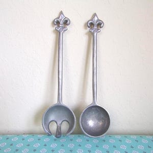 Puede incluir: Un par de utensilios de metal plateado para servir ensalada con mangos largos y delgados rematados con un emblema de flor de lis. Un utensilio es una cuchara de cuenco redondo y el otro es una cuchara ranurada con tres puntas en la base.