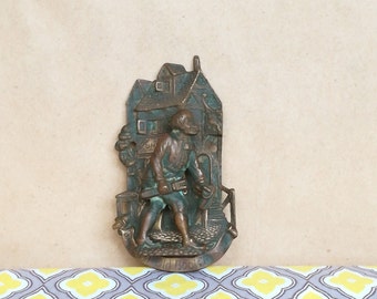May include: A bronze door knocker depicting a man in a medieval-style outfit holding a hammer and standing in front of a building with the text "Old Bocks" on the bottom.