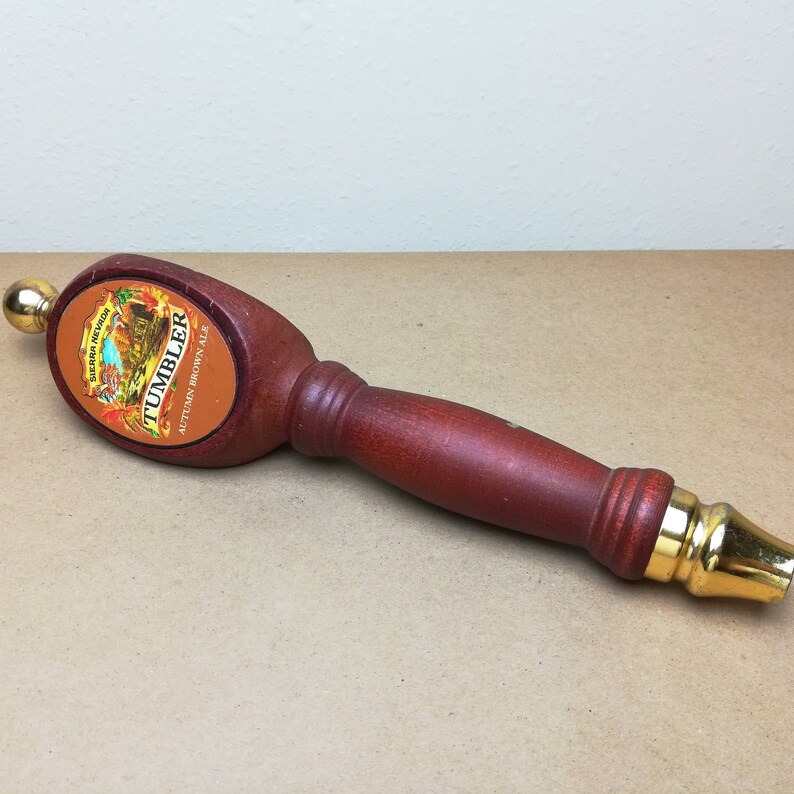 May include: A wooden beer tap handle with a dark reddish-brown finish and gold-colored metal accents at both ends. The oval-shaped head features a label for Sierra Nevada Tumbler Autumn Brown Ale with an illustration of autumn leaves and trees.