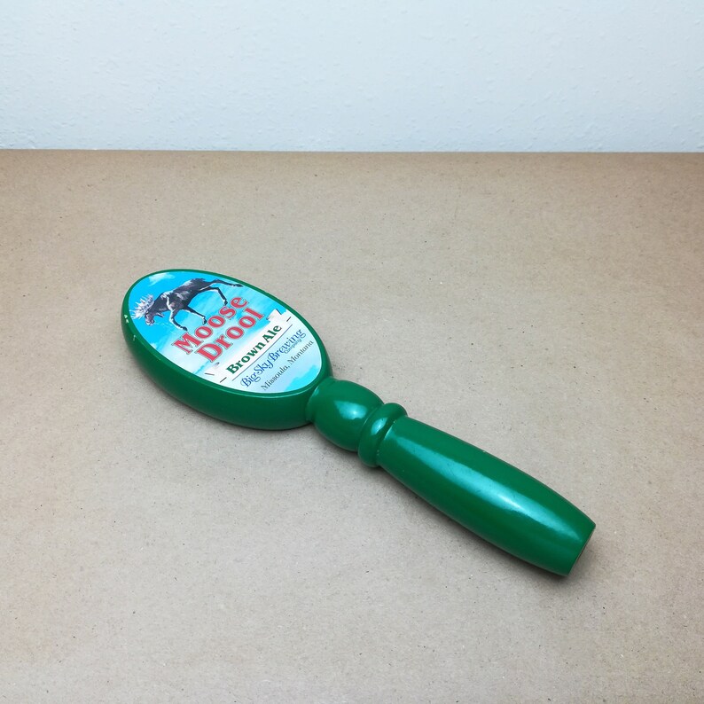 May include: A green beer tap handle with an oval top featuring a graphic of a moose and the text Moose Drool Brown Ale Big Sky Brewing Missoula, Montana. The handle has a smooth, glossy finish and a rounded, ergonomic grip for easy operation.