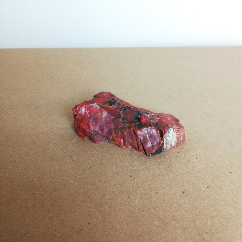 May include: A red, irregularly shaped piece of red jasper with white and black veins. The stone is sitting on a brown surface.