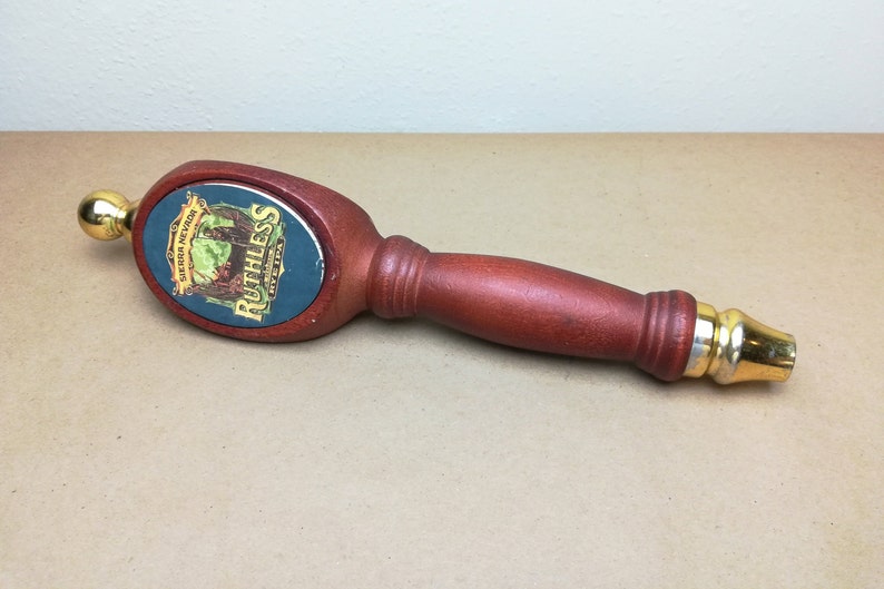 May include: A wooden beer tap handle with a dark reddish-brown finish and gold-colored metal accents at the top and bottom. The oval center features a label with the text Sierra Nevada Ruthless Rye IPA and an illustration of a figure in a landscape.