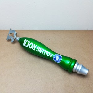 May include: A green Rolling Rock beer tap handle featuring a metallic silver finish at the top and bottom. The body is glossy green with the white text ROLLING ROCK printed along the side and a circular blue and white logo featuring a horse head.