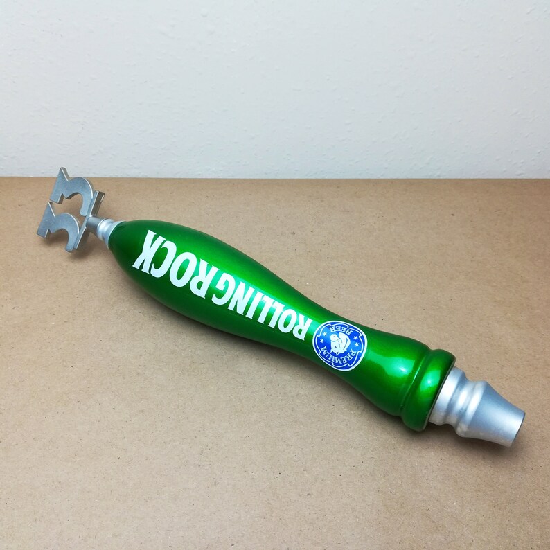 May include: A green Rolling Rock beer tap handle featuring a metallic silver finish at the top and bottom. The body is glossy green with the white text ROLLING ROCK printed along the side and a circular blue and white logo featuring a horse head.