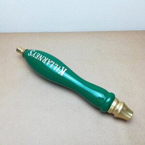 May include: A glossy green beer tap handle featuring the white text KILLARNEY'S printed along the side. The handle has a smooth, elongated, rounded shape with metallic gold-colored fittings at both the top and bottom ends of the object.