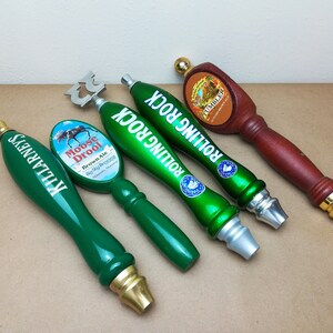 May include: Four beer tap handles arranged in a row. One is green with Killarney's text, one is green with a Moose Drool Brown Ale label, two are green with Rolling Rock text, and one is a brown wooden handle with a Tumbler Autumn Brown Ale label.