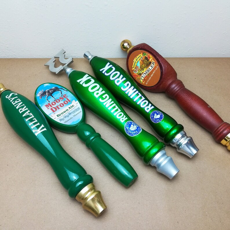 May include: Four beer tap handles arranged in a row. One is green with Killarney's text, one is green with a Moose Drool Brown Ale label, two are green with Rolling Rock text, and one is a brown wooden handle with a Tumbler Autumn Brown Ale label.
