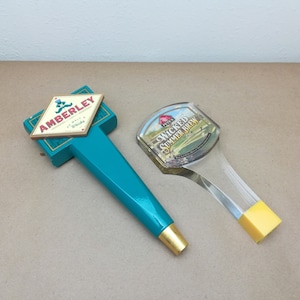 May include: Two beer tap handles. One is a teal, T-shaped handle with a diamond-shaped plaque reading AMBERLEY MALT WHISKY and a gold-colored base. The other is a clear, curved handle with a label reading PETE'S WICKED SUMMER BREW and a yellow base.