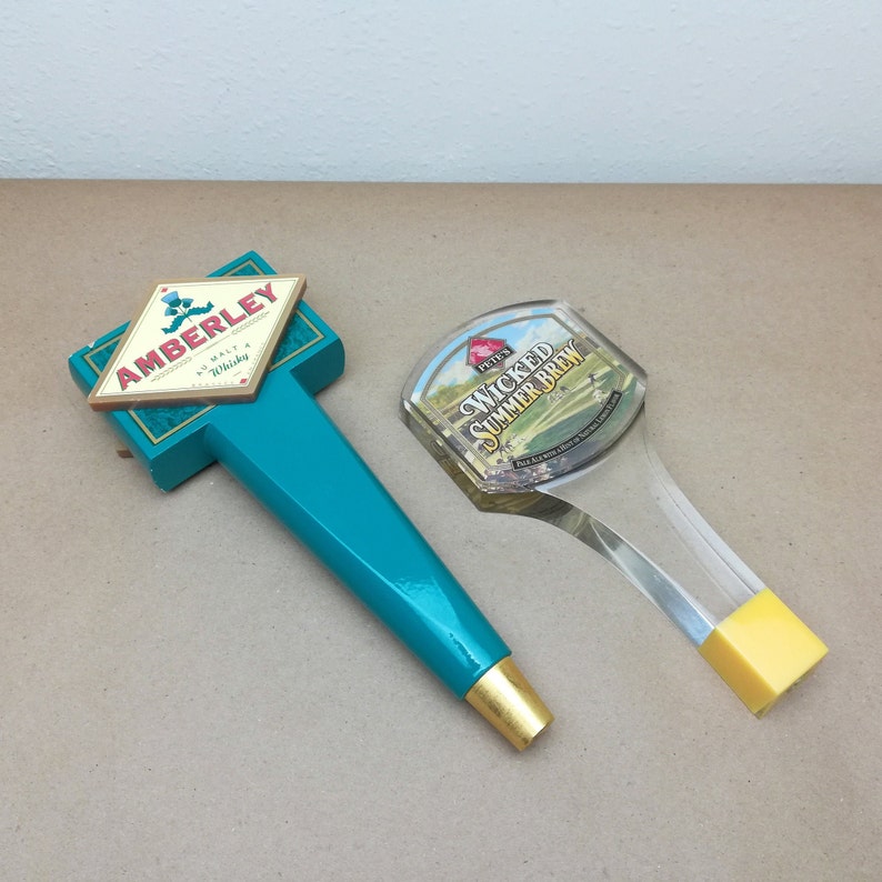 May include: Two beer tap handles. One is a teal, T-shaped handle with a diamond-shaped plaque reading AMBERLEY MALT WHISKY and a gold-colored base. The other is a clear, curved handle with a label reading PETE'S WICKED SUMMER BREW and a yellow base.