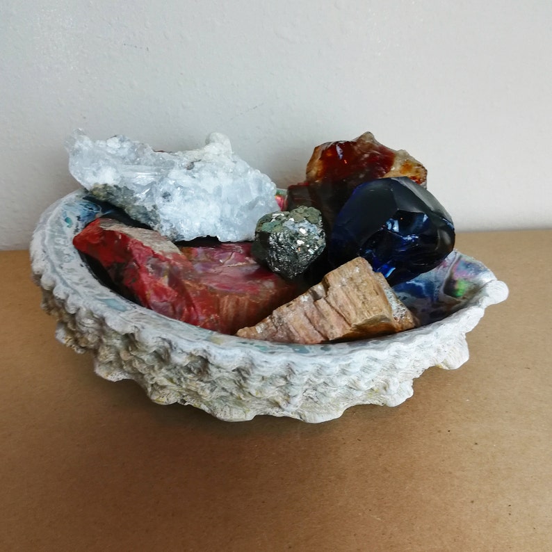 May include: A collection of colorful crystals and stones, including a blue crystal, a red and white crystal, a brown and gold crystal, and a white crystal, are displayed in a white, textured bowl.