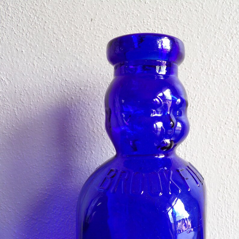 May include: A cobalt blue glass bottle with a rounded, human-like face. The bottle has a wide mouth and a rounded body. The word "BROOKLYN" is embossed on the front of the bottle.