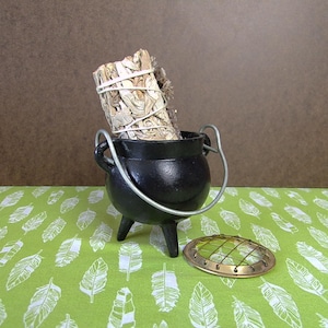 May include: A black cast iron cauldron with a wire handle, sitting on a green tablecloth with white feather designs. A bundle of white sage is inside the cauldron, and a brass mesh incense burner sits on the tablecloth.