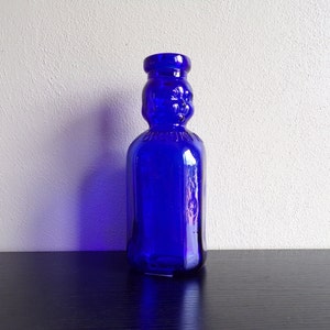May include: A cobalt blue glass bottle with a unique design. The bottle has a rounded body and a narrow neck with a raised, embossed design.