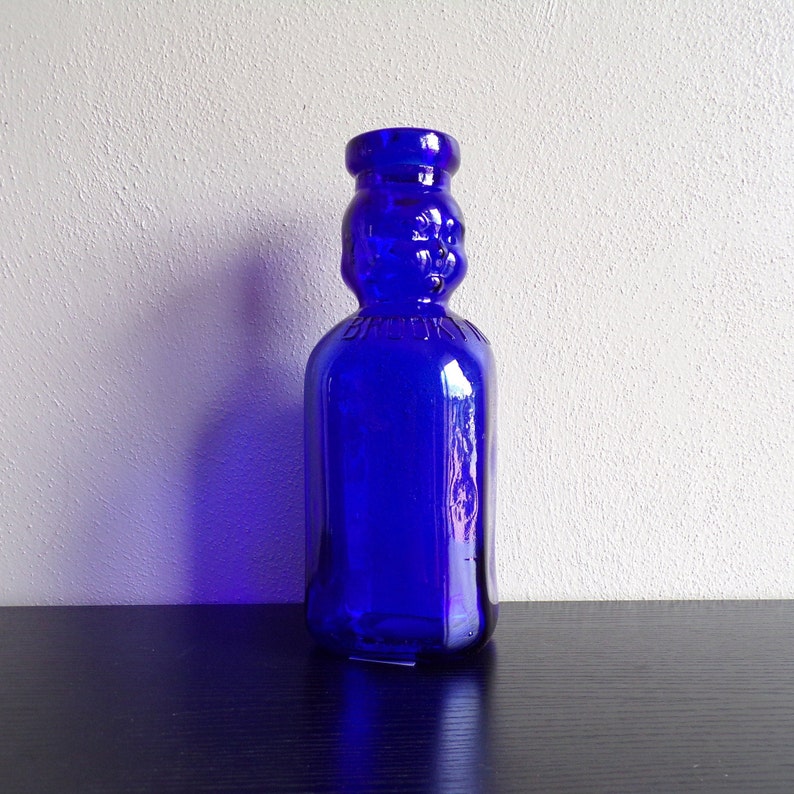 May include: A cobalt blue glass bottle with a unique design. The bottle has a rounded body and a narrow neck with a raised, embossed design.