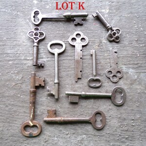 May include: A collection of ten antique keys, some with intricate designs, including a key with the words "USA" and "STAINLESS" on it. The keys are arranged on a gray textured surface. LOT K.