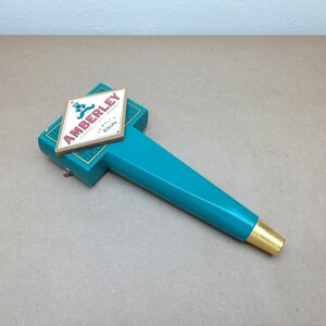 May include: A teal beer tap handle featuring a diamond-shaped plaque with the text AMBERLEY AU MALT A WHISKY and a small blue thistle graphic. The handle has a smooth, glossy finish and a gold-colored metal ferrule at the bottom end.