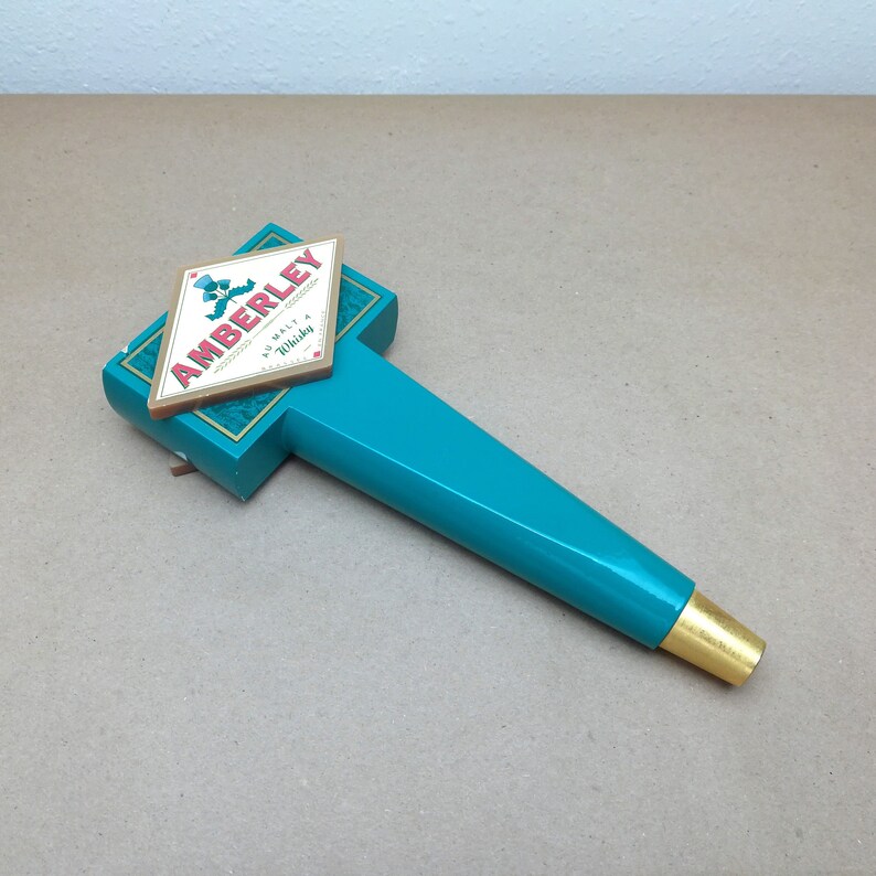May include: A teal beer tap handle featuring a diamond-shaped plaque with the text AMBERLEY AU MALT A WHISKY and a small blue thistle graphic. The handle has a smooth, glossy finish and a gold-colored metal ferrule at the bottom end.
