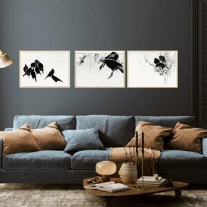 May include: Three framed art prints featuring black bird illustrations on a white background. The artwork is displayed above a blue sofa with brown and blue pillows. The room has a dark gray wall and a gold floor lamp.