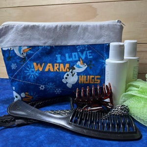 May include: A blue quilted zippered pouch with a white trim and a snowman design. The pouch has the text "I love warm hugs" on it. The pouch is surrounded by various items, including two bottles of lotion, a green bath sponge, and two hair clips.