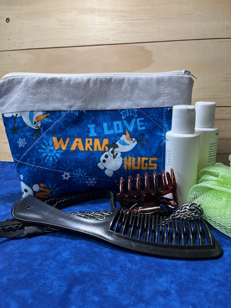 May include: A blue quilted zippered pouch with a white trim and a snowman design. The pouch has the text "I love warm hugs" on it. The pouch is surrounded by various items, including two bottles of lotion, a green bath sponge, and two hair clips.