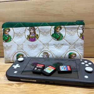 May include: A white zippered pouch with a green trim and a repeating pattern of the Legend of Zelda characters Link and Princess Zelda. The pouch is lying on a wooden surface next to a gray Nintendo Switch console and three game cartridges.