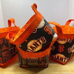 May include: Three fabric baskets with orange handles and trim. The baskets feature a black background with baseballs and the San Francisco Giants logo in orange and white. The baskets are stacked and arranged on a wooden surface.