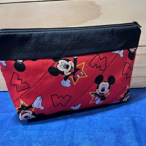 Puede incluir: Bolsa roja con cremallera y solapa superior negra. La bolsa est&aacute; decorada con un patr&oacute;n repetido de Mickey Mouse y estrellas amarillas.