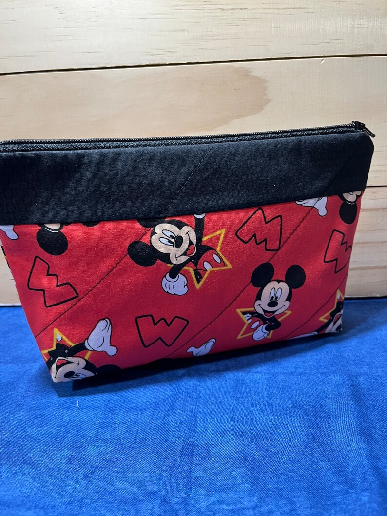 Puede incluir: Bolsa roja con cremallera y solapa superior negra. La bolsa est&aacute; decorada con un patr&oacute;n repetido de Mickey Mouse y estrellas amarillas.