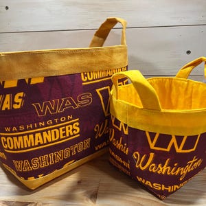 May include: Two fabric storage bins with yellow handles. The larger bin is burgundy with yellow trim and the text "WAS WASHINGTON COMMANDERS WASHINGTON" printed on it. The smaller bin is burgundy with yellow trim and the text "Washington WASHINGTON" printed on it.