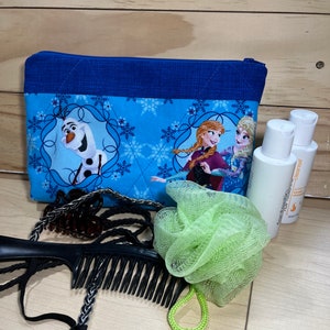 May include: A blue zippered pouch with a Frozen theme featuring Olaf, Anna, and Elsa. The pouch is on a wooden surface with a green bath sponge, a black comb, and two bottles of body wash.