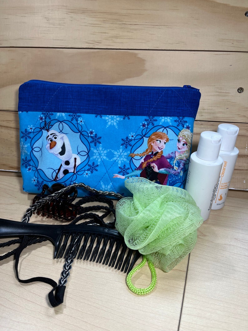 May include: A blue zippered pouch with a Frozen theme featuring Olaf, Anna, and Elsa. The pouch is on a wooden surface with a green bath sponge, a black comb, and two bottles of body wash.