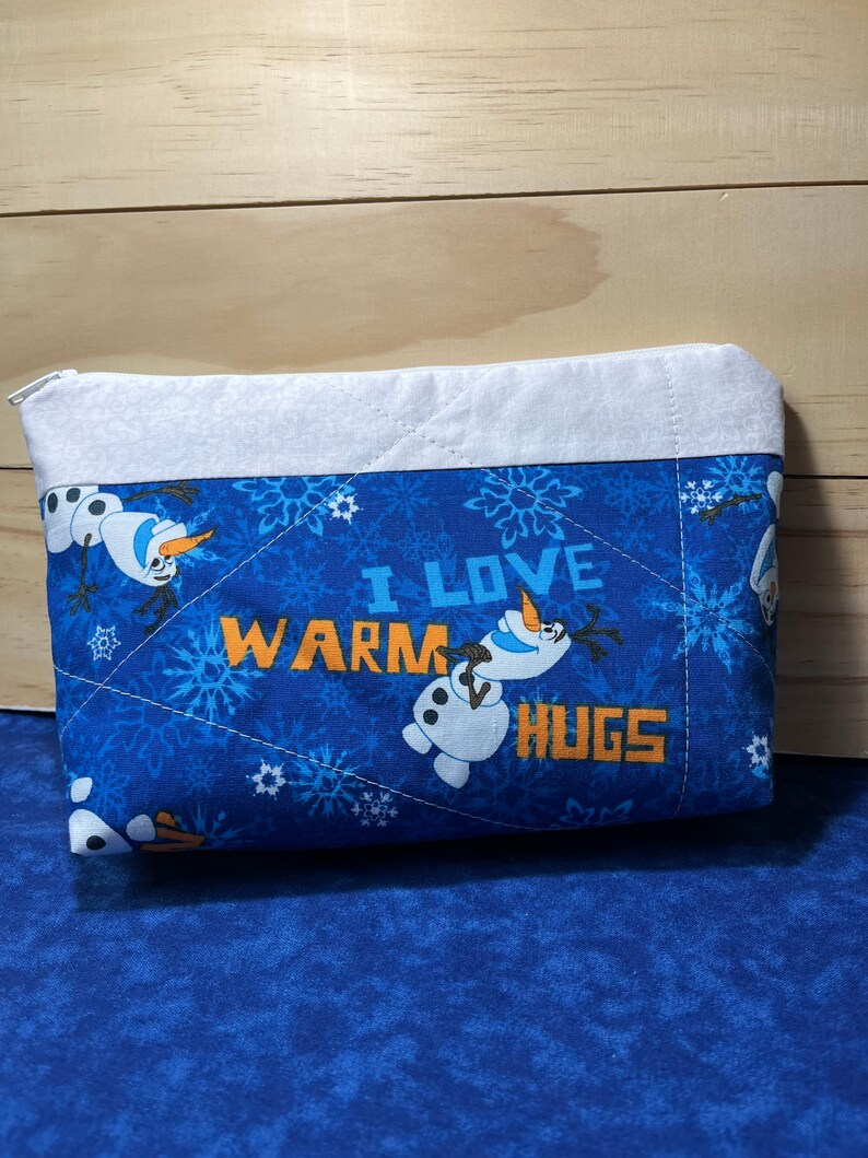 May include: Blue quilted zippered pouch with a white border. The pouch features a snowman design with the text "I love warm hugs".