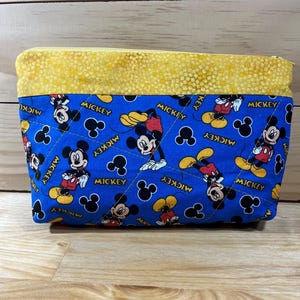 Puede incluir: Una bolsa azul con cremallera y parte superior amarilla. La bolsa est&aacute; decorada con un patr&oacute;n repetido de Mickey Mouse en rojo, amarillo y negro. Las palabras "Mickey" est&aacute;n impresas en la bolsa.
