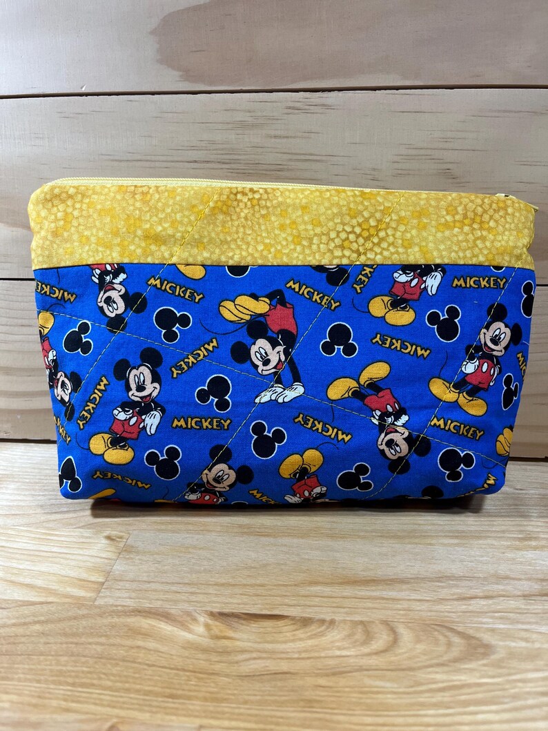Puede incluir: Una bolsa azul con cremallera y parte superior amarilla. La bolsa est&aacute; decorada con un patr&oacute;n repetido de Mickey Mouse en rojo, amarillo y negro. Las palabras "Mickey" est&aacute;n impresas en la bolsa.