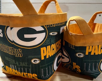 May include: Two green and gold fabric storage bins with the Green Bay Packers logo and team colors. The bins have a handle on the top and are made of a sturdy fabric.