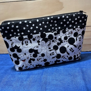 Puede incluir: Una bolsa con cremallera negra y blanca con un estampado de Mickey Mouse. La bolsa tiene un ribete de tela de lunares negros y blancos.