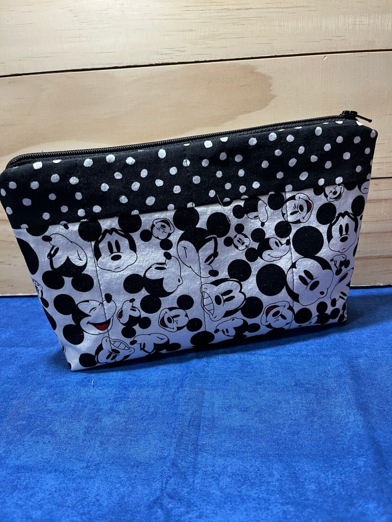 Puede incluir: Una bolsa con cremallera negra y blanca con un estampado de Mickey Mouse. La bolsa tiene un ribete de tela de lunares negros y blancos.