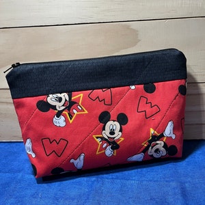 Puede incluir: Bolsa de cremallera acolchada roja con cremallera negra y ribete de tela negra. La bolsa presenta un patr&oacute;n repetido de Mickey Mouse en estrellas amarillas sobre un fondo rojo.