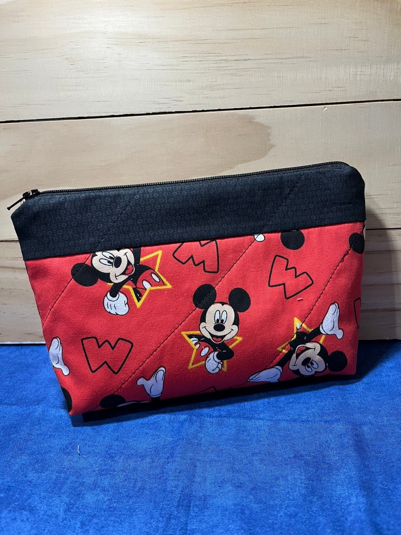 Puede incluir: Bolsa de cremallera acolchada roja con cremallera negra y ribete de tela negra. La bolsa presenta un patr&oacute;n repetido de Mickey Mouse en estrellas amarillas sobre un fondo rojo.