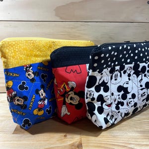 Puede incluir: Tres estuches con cremallera con diferentes patrones de Mickey Mouse. El estuche de la izquierda es azul con detalles amarillos y presenta a Mickey Mouse en varias poses. El estuche del medio es rojo con detalles negros y presenta a Mickey Mouse con una estrella roja. El estuche de la derecha es blanco con detalles negros y presenta la cabeza de Mickey Mouse en varias poses.