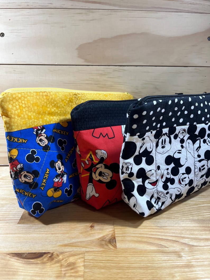 Puede incluir: Tres estuches con cremallera con diferentes patrones de Mickey Mouse. El estuche de la izquierda es azul con detalles amarillos y presenta a Mickey Mouse en varias poses. El estuche del medio es rojo con detalles negros y presenta a Mickey Mouse con una estrella roja. El estuche de la derecha es blanco con detalles negros y presenta la cabeza de Mickey Mouse en varias poses.