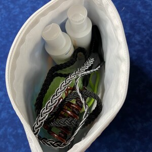 May include: A white zippered pouch containing two clear bottles of lotion, a black and silver braided hair tie, and a brown hair clip.