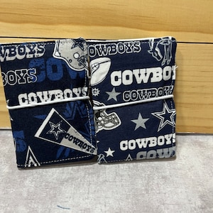 May include: Two blue and white fabric pouches with a Dallas Cowboys theme. The pouches have a variety of Cowboys logos and designs, including the team's star logo, a football, and the word "Cowboys".