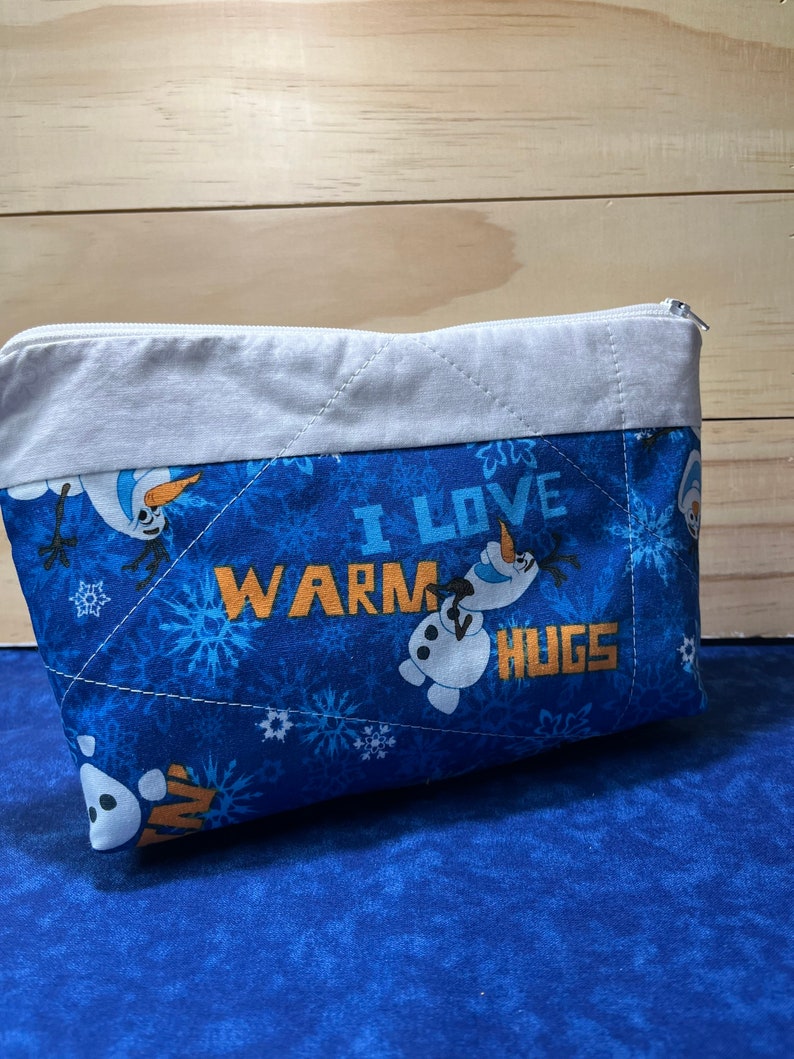 May include: Blue quilted zippered pouch with a white trim. The pouch features a snowman design and the text "I love warm hugs".
