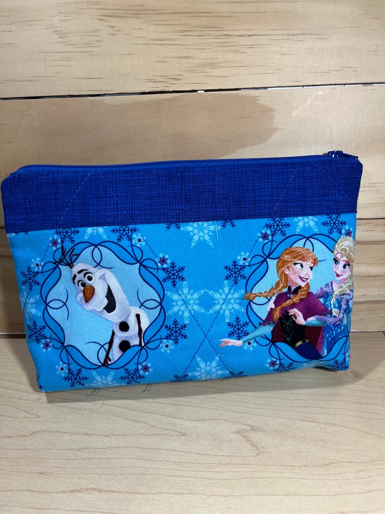 May include: A blue zippered pouch with a quilted design. The pouch features a print of Elsa, Anna, and Olaf from the movie Frozen. The print is on a light blue background with white snowflakes.