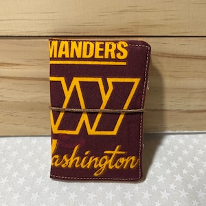 May include: A burgundy fabric wallet with a gold embroidered Washington Commanders logo. The logo features the team's 'W' and the words 'Commanders' and 'Washington'.