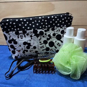 Puede incluir: Una bolsa con cremallera de lunares negros y blancos con un estampado de Mickey Mouse. La bolsa est&aacute; llena de art&iacute;culos de tocador, incluyendo dos botellas blancas, una esponja de ba&ntilde;o verde, una diadema negra y una pinza para el pelo marr&oacute;n.