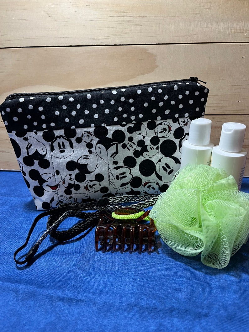 Puede incluir: Una bolsa con cremallera de lunares negros y blancos con un estampado de Mickey Mouse. La bolsa est&aacute; llena de art&iacute;culos de tocador, incluyendo dos botellas blancas, una esponja de ba&ntilde;o verde, una diadema negra y una pinza para el pelo marr&oacute;n.