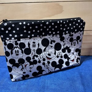 Puede incluir: Una bolsa con cremallera negra y blanca con un estampado de Mickey Mouse. La bolsa tiene un ribete de tela de lunares negros y blancos.